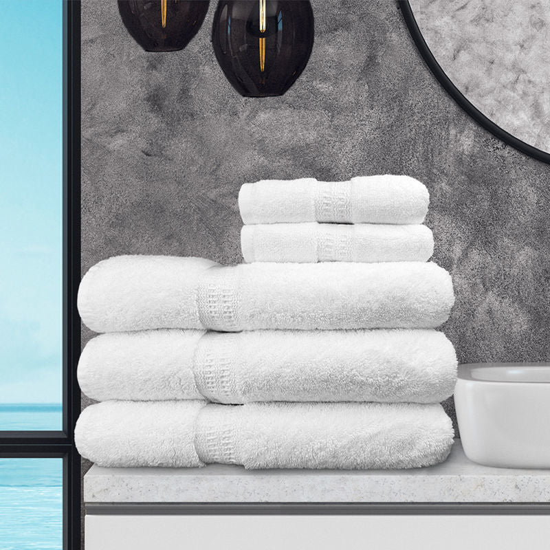 A stack of white Ganesh Mills Oxford Reserve Hand Towels sits on a bathroom countertop by a white sink, with a round mirror, two dark hanging lights, and a gray textured wall. A window shows blue sky and water.