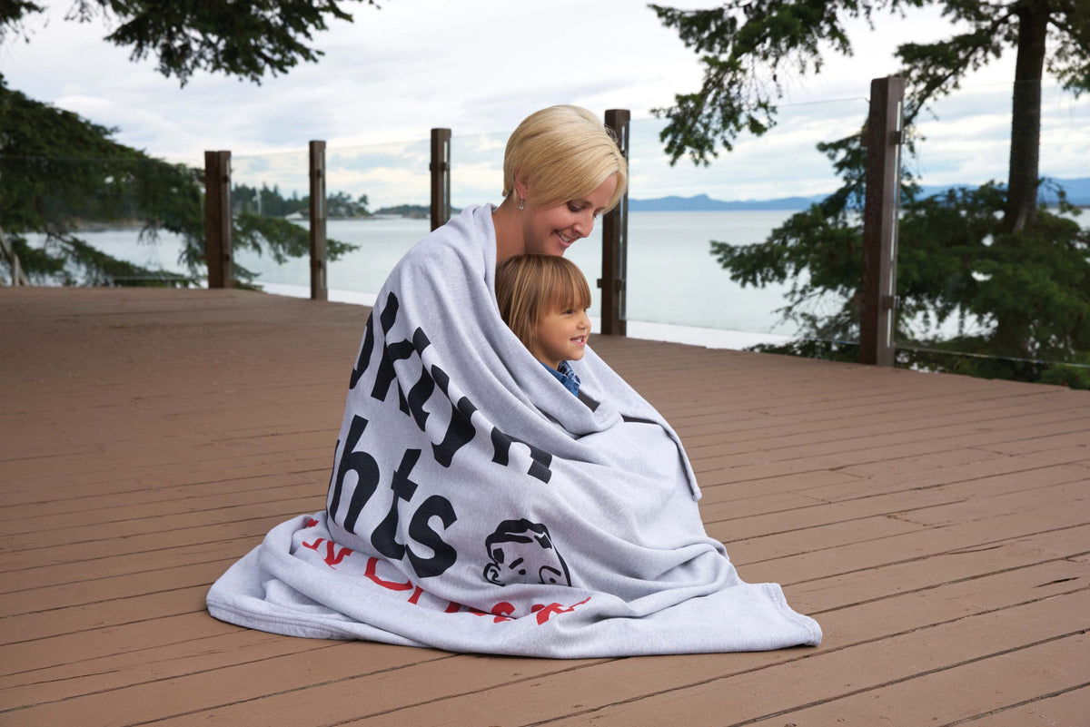 A woman and child snuggle in a Pro Towels Oversized Sweatshirt Blanket on a wooden deck by a lake, surrounded by trees with mountains in the background.