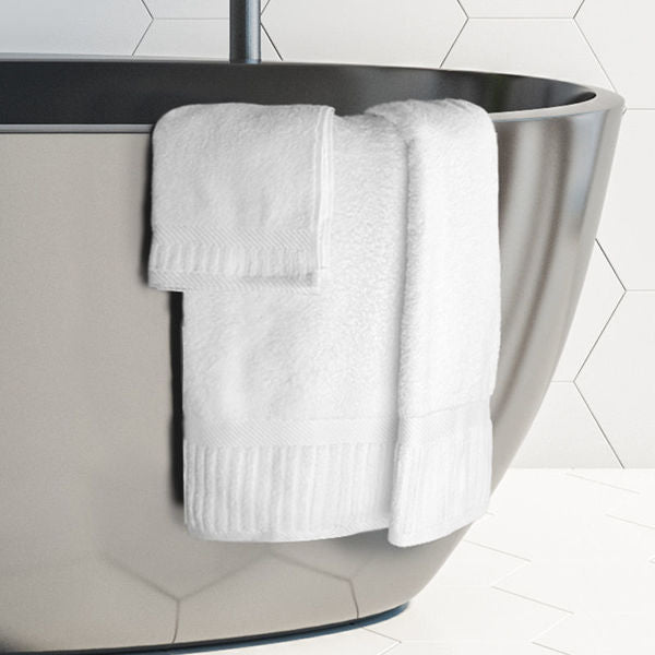 A white Oxford Signature bath towel by Ganesh Mills is neatly draped over a modern gray bathtub, matched with a washcloth. White hexagon tiles form the backdrop for this ringspun cotton towel set.
