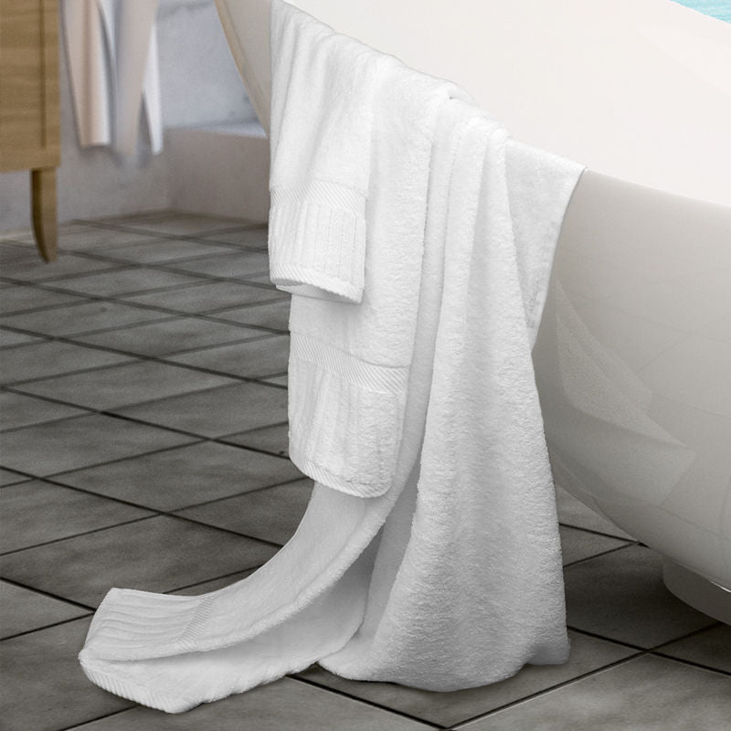 The Ganesh Mills Bathmat - White Oxford Signature Towel, made of ringspun cotton, is draped over a white bathtub in a tiled bathroom, with part of the bathmat hanging down to touch the floor.
