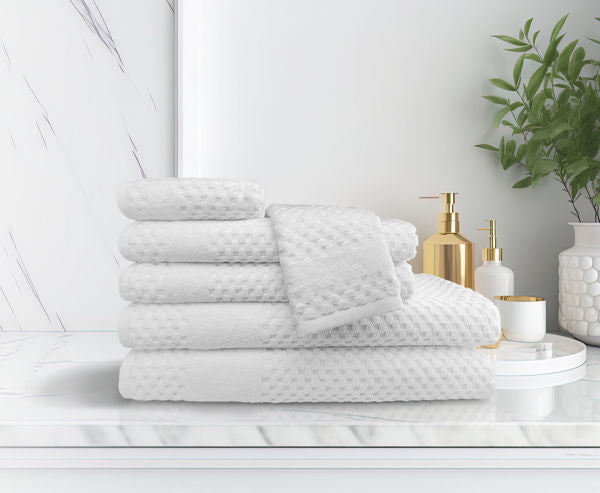 A white Bath Mat from the Ganesh Mills Oxford Tuscany Towel Collection rests on a marble bathroom counter beside a gold soap dispenser, a white container, and a green potted plant, with a marble wall in the background.