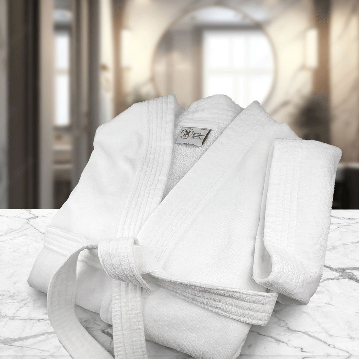 A Ganesh Mills Oxford Velour Bathrobe and towel are arranged on white marble, complemented by a White Replacement Belt for Oxford Velour Bathrobes, creating a luxurious display with an elegantly blurred interior in the background.