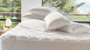 A stack of white pillows sits on a neatly made bed fitted with the Ganesh Mills Oxford Quilted Bed Pads, next to a large window overlooking trees.