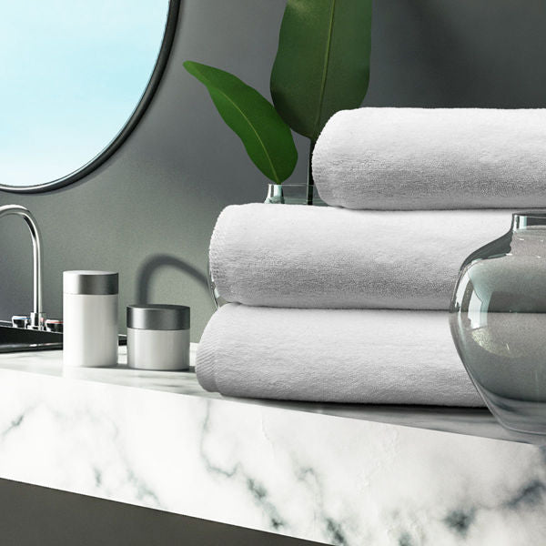 A modern bathroom with a marble countertop, glass vases, a plant, and stacked Bath Sheet / Pool Towels – White Oxford Platinum Towel by Ganesh Mills, all bathed in soft natural light beside a round mirror.