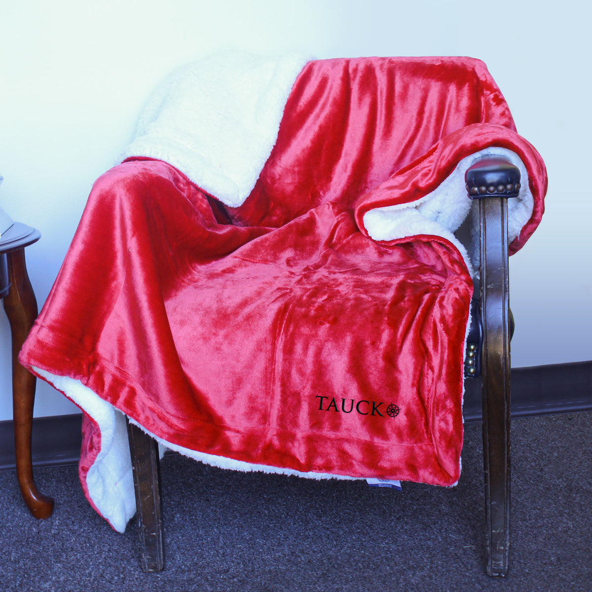 A cozy red and white Palmetto Challenger Blanket by Pro Towels, custom embroidered with "TAUCK," is draped over a brown upholstered chair against a white wall.