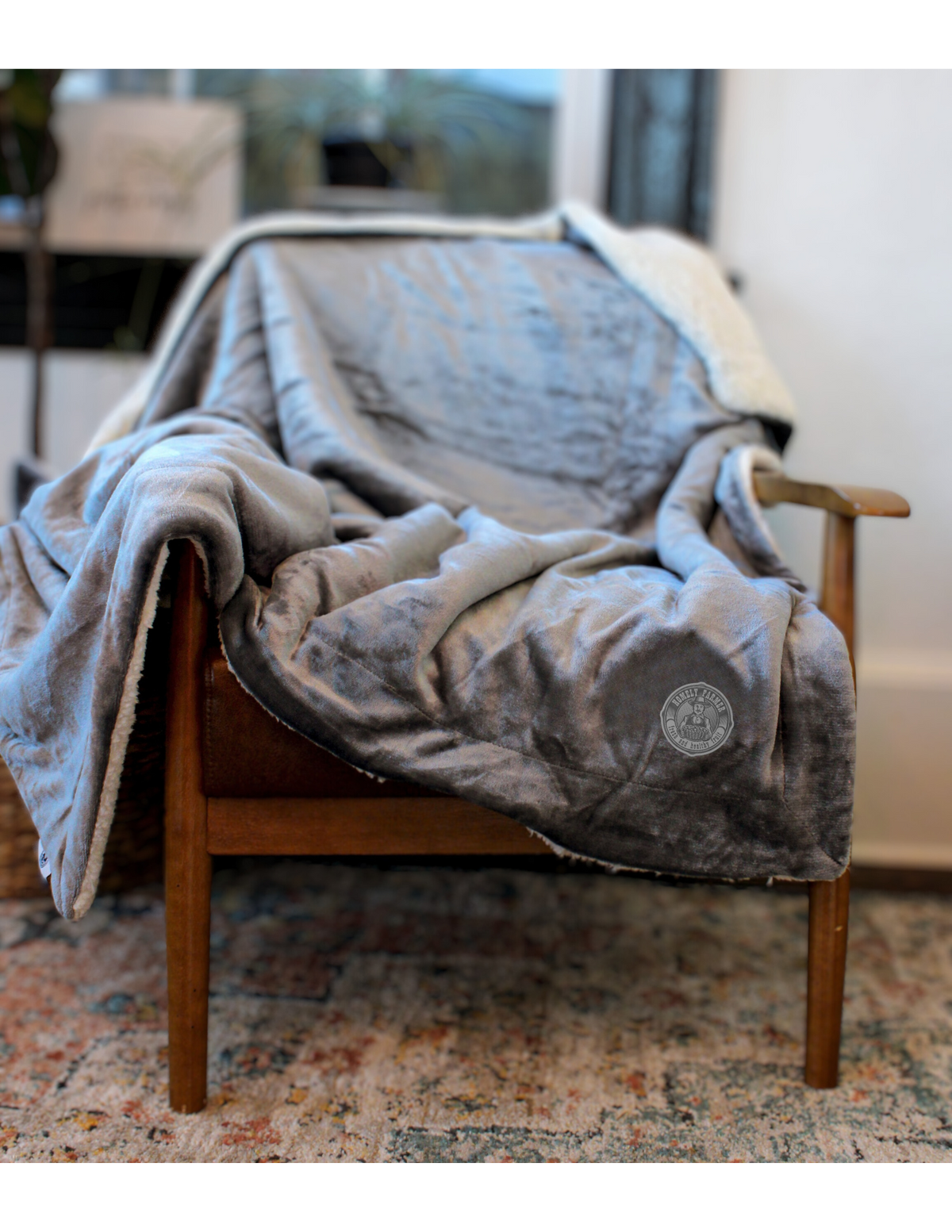 The Palmetto Challenger Blanket by Pro Towels, in cozy gray with a custom round patch, is draped over a wooden armchair in a warmly lit room featuring a patterned rug.