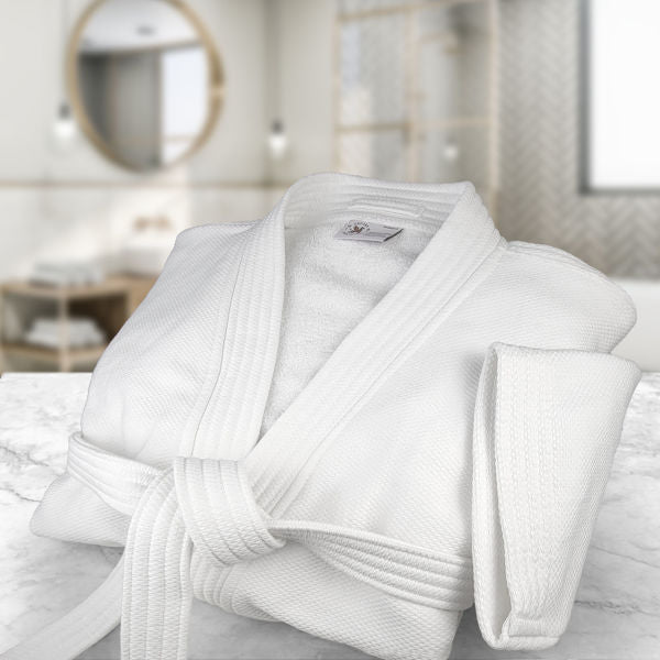 The Ganesh Mills Kimono - White Oxford Terry Bathrobe with belt is neatly folded on a marble surface in a modern, softly lit bathroom featuring a round mirror and blurred background.