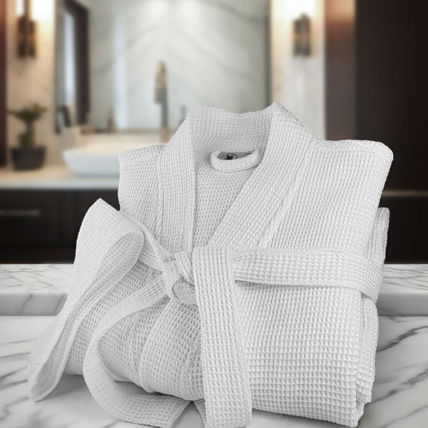 A folded Oxford Honeycomb Waffle Bathrobe in white by Ganesh Mills with belt sits on a marble countertop in a modern bathroom—perfect for those seeking bulk spa bathrobes.