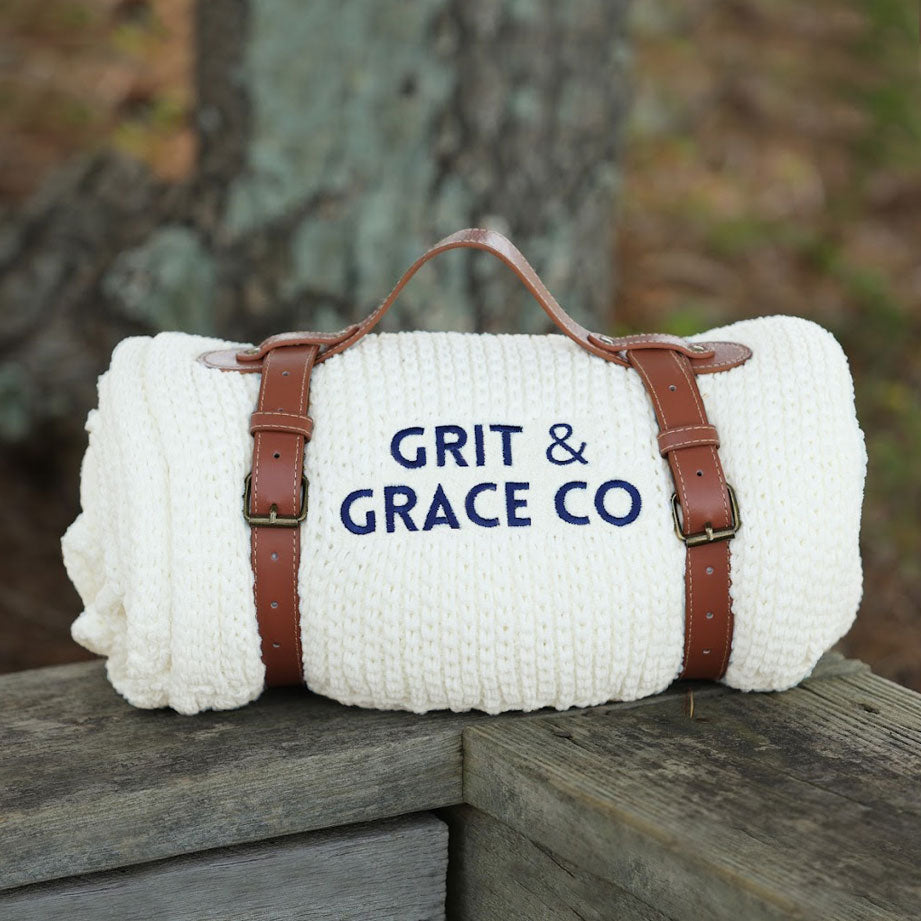 The Riviera Throw EnviroFibr by Pro Towels, a white custom embroidered blanket with brown leather straps and handle featuring GRIT & GRACE CO, rests outdoors on wood—an eco-friendly throw ideal for cozy gatherings.