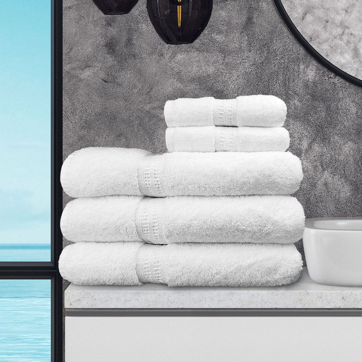 A stack of Ganesh Mills Bath Sheet / Pool Towel - White Oxford Reserve Towel is arranged on a bathroom counter by a white sink, set against a gray textured wall, round mirror, and window overlooking the blue ocean.