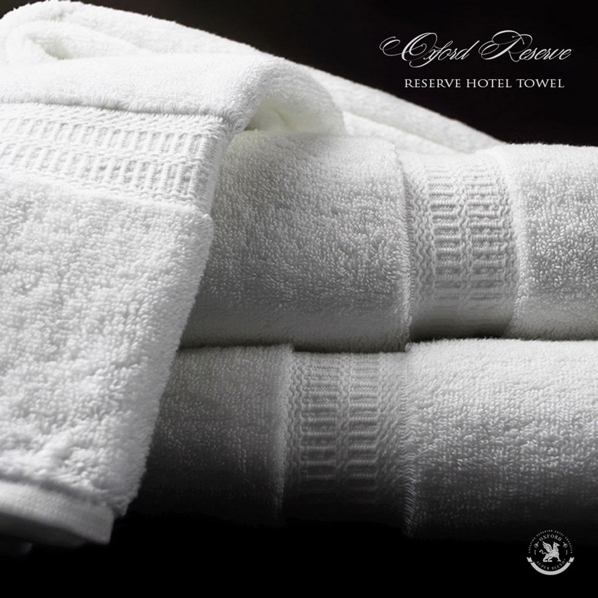 Close-up of three neatly folded, plush white Ganesh Mills Hand Towels from the Oxford Reserve Towel Collection stacked on a dark surface, featuring soft, spa-quality texture with decorative bands. Text reads: Oxford Reserve Hand Towel.