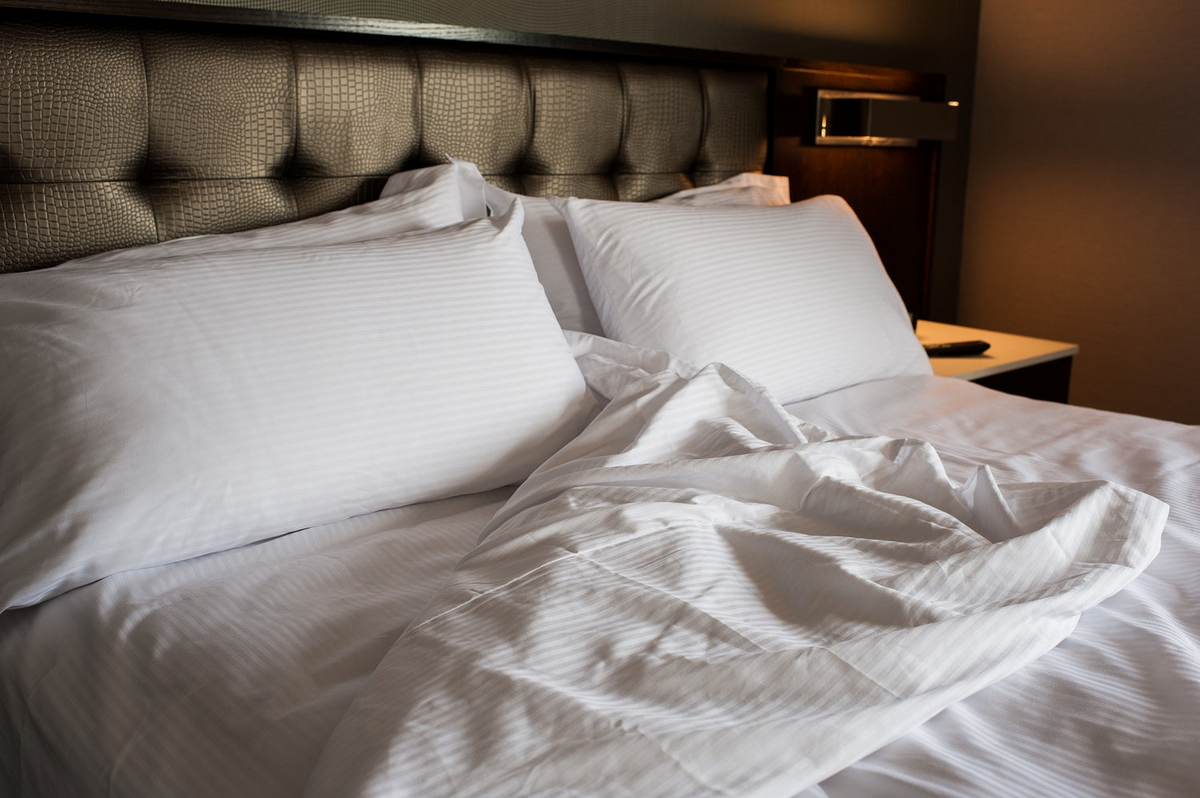 A neatly made bed with a tufted headboard, white pillows, and Thomaston Mills Duvet Covers—310 Thread Count Satin Stripe—partially pulled back. A bedside table with a phone sits nearby under warm, soft lighting that highlights quality hospitality bedding.
