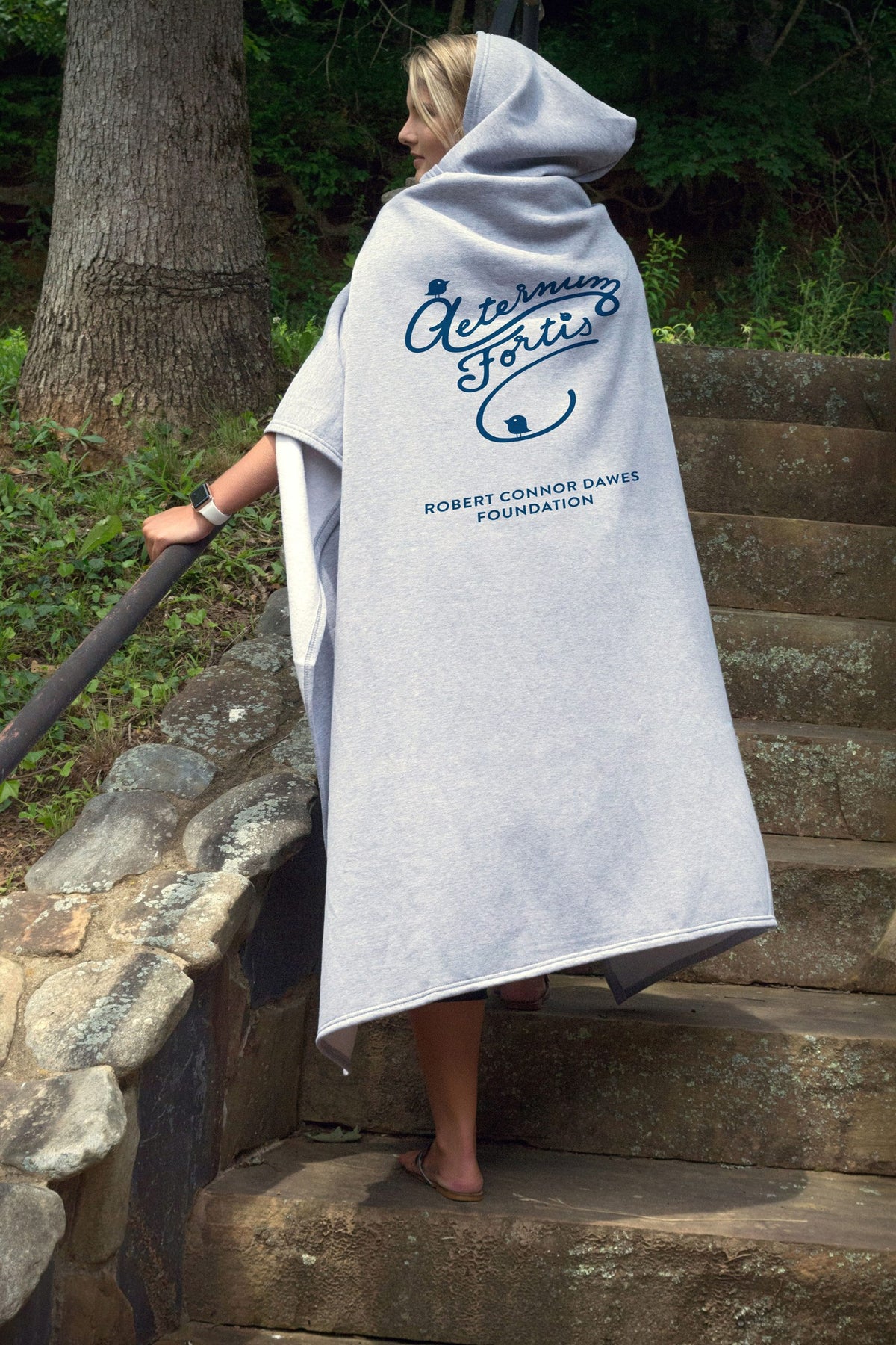 A person in a light gray Pro Towels Sweatshirt Sport Hoodie with custom Aeternum Fortis and Robert Connor Dawes Foundation embroidery on the back stands outdoors on stone steps, holding the railing and facing away.