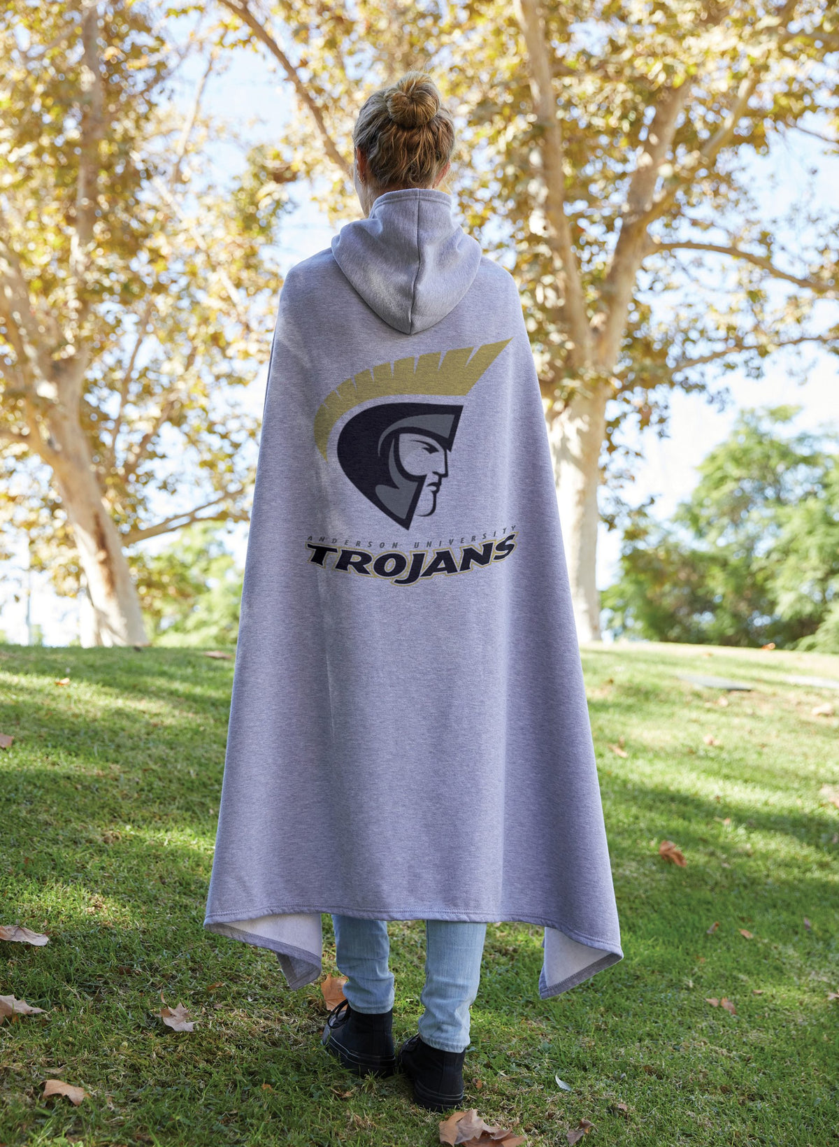 Outdoors on grass, a person faces away wearing the Pro Towels Sweatshirt Sport Hoodie in gray, featuring a large Trojans logo on the back. Sunlit trees with golden leaves fill the background.