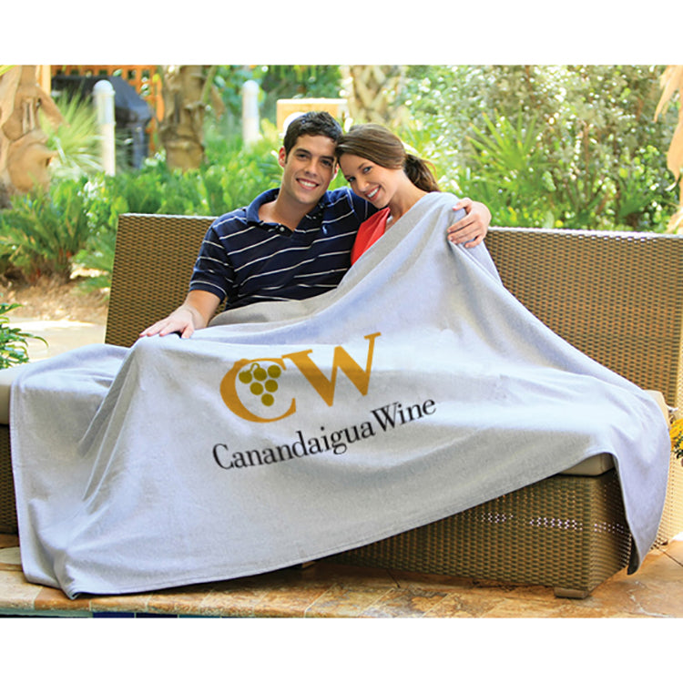 A smiling couple sits together on a wicker outdoor sofa, wrapped in the Pro Towels Oversized Sweatshirt Blanket. Lush greenery in the background highlights this cozy and stylish blanket, perfect for relaxing outdoors.
