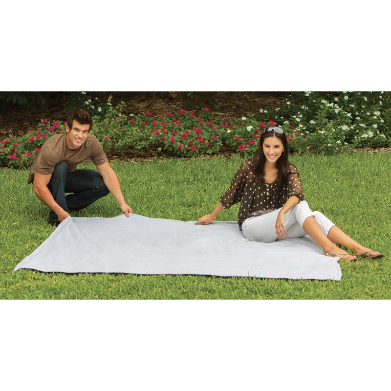 A man and a woman are laying out the Pro Towels Oversized Sweatshirt Blanket on green grass in a garden, with pink and white flowers in the background.