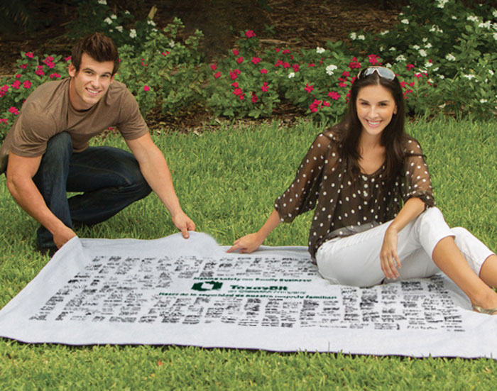 A man and woman smile while sitting on the grass, spreading out the Pro Towels Oversized Sweatshirt Blanket with black handwritten text and a green logo. Blooming pink and white flowers and bushes can be seen in the background.