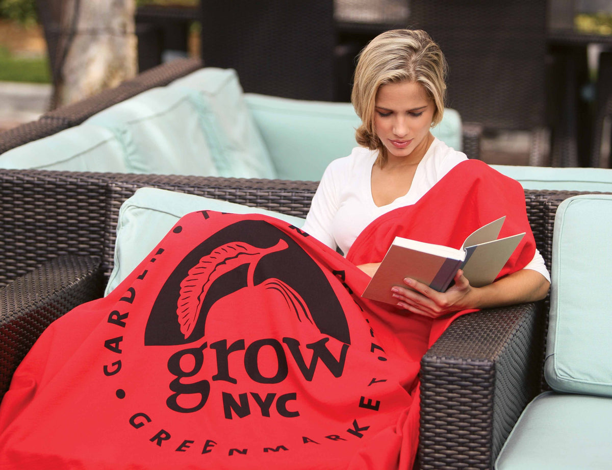 A woman sits on an outdoor wicker sofa with light blue cushions, wrapped in a Pro Towels Sweatshirt Blanket with a "grow NYC Greenmarket" logo, as she reads a book.