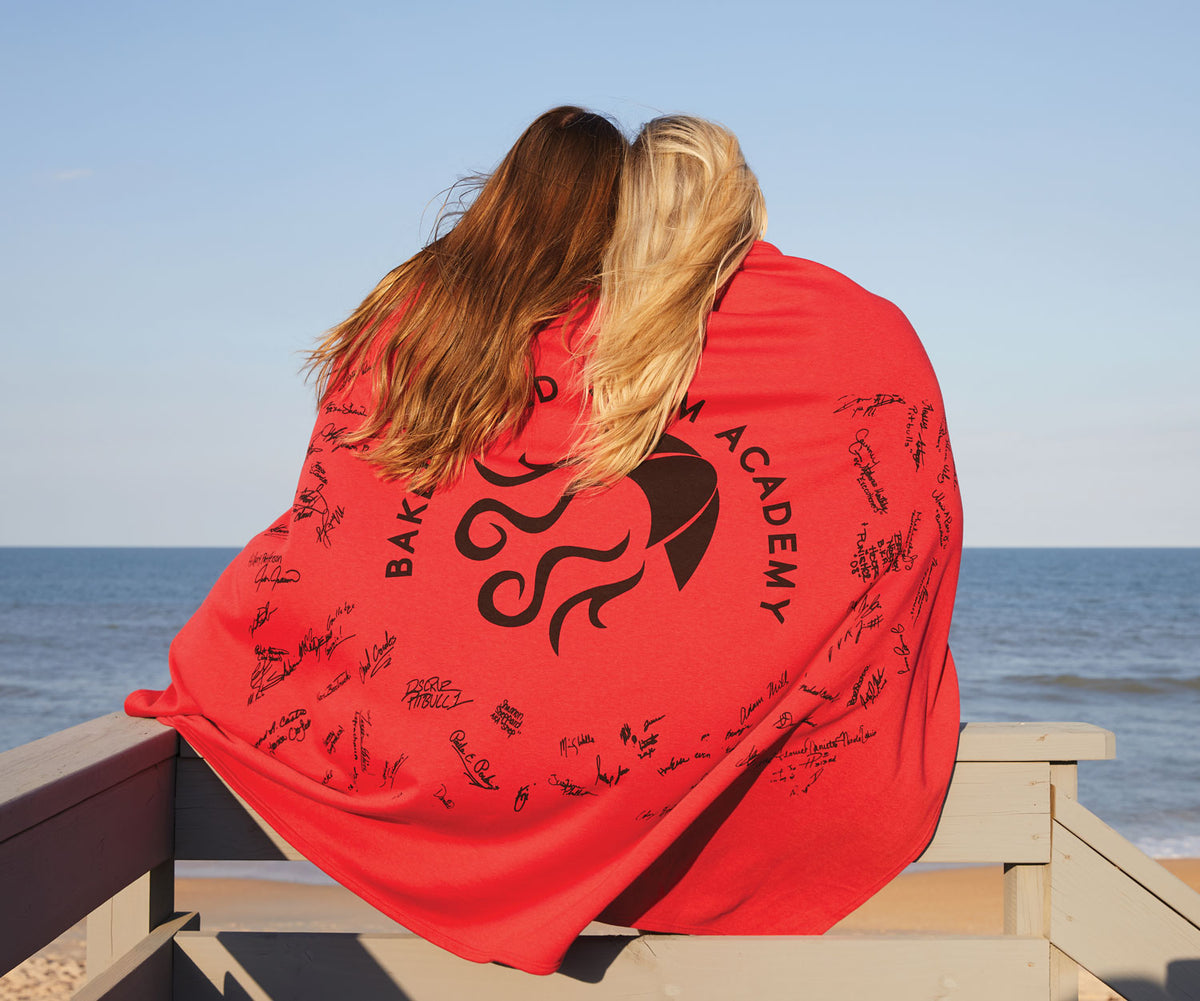 Two people sit on a wooden railing by the ocean, wrapped together in a red Pro Towels Sweatshirt Blanket with black signatures. With their backs to the camera, they gaze at the sea under a clear blue sky.