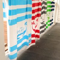 Sandbar Stripe Beach Towels by Pro Towels in blue, red, gray, and green hang side by side. Each towel shows a white whale design with “THE ISLAND” text, all displayed on a wooden deck.