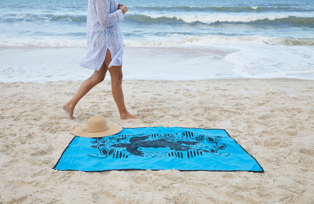 A person in a white cover-up walks barefoot on the sand near a Pro Towels Sand Repellent Beach Blanket and a straw hat, with ocean waves in the background.