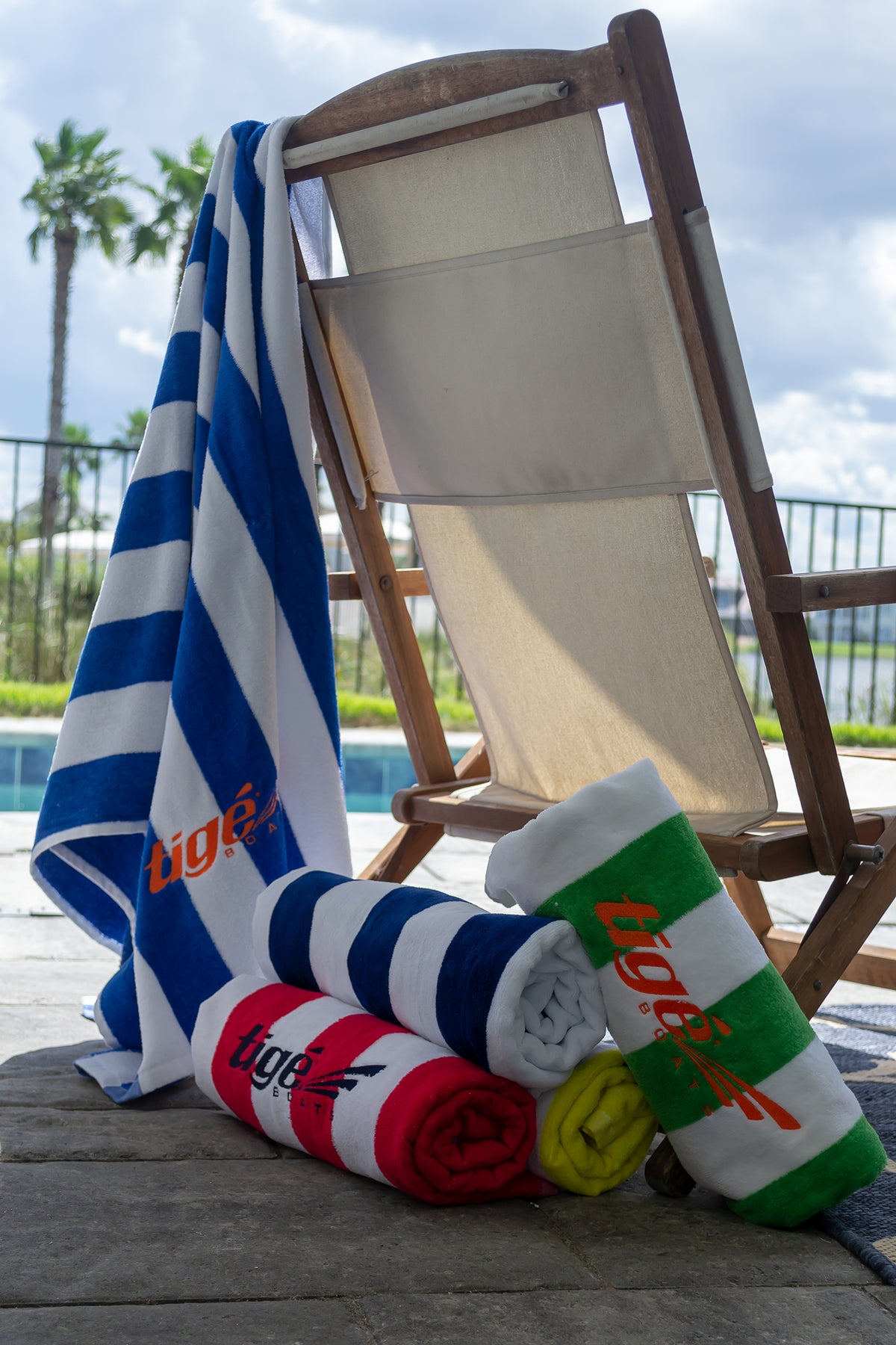 A poolside lounge chair displays a Pro Towels Standard Cabana Beach Towel in blue and white stripes, along with four rolled towels—red, green, yellow, and blue—each labeled "tige," set against a backdrop of a pool and palm trees.