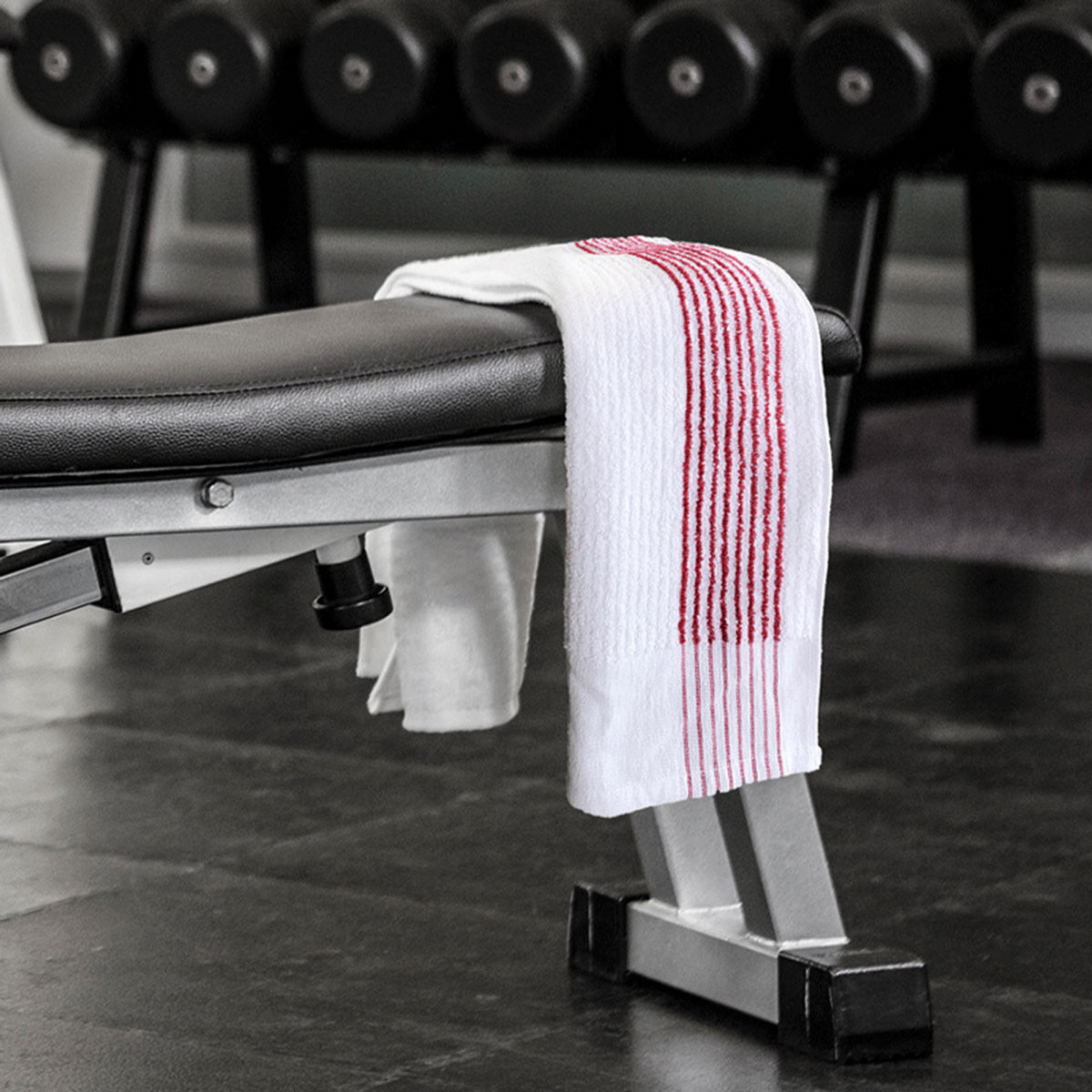 The Ganesh Mills Oxford Super Gym Towel—white with 8 color stripes—is draped over a gym bench’s backrest. Behind, black dumbbells line a rack, and the spotless dark gym floor completes the scene.