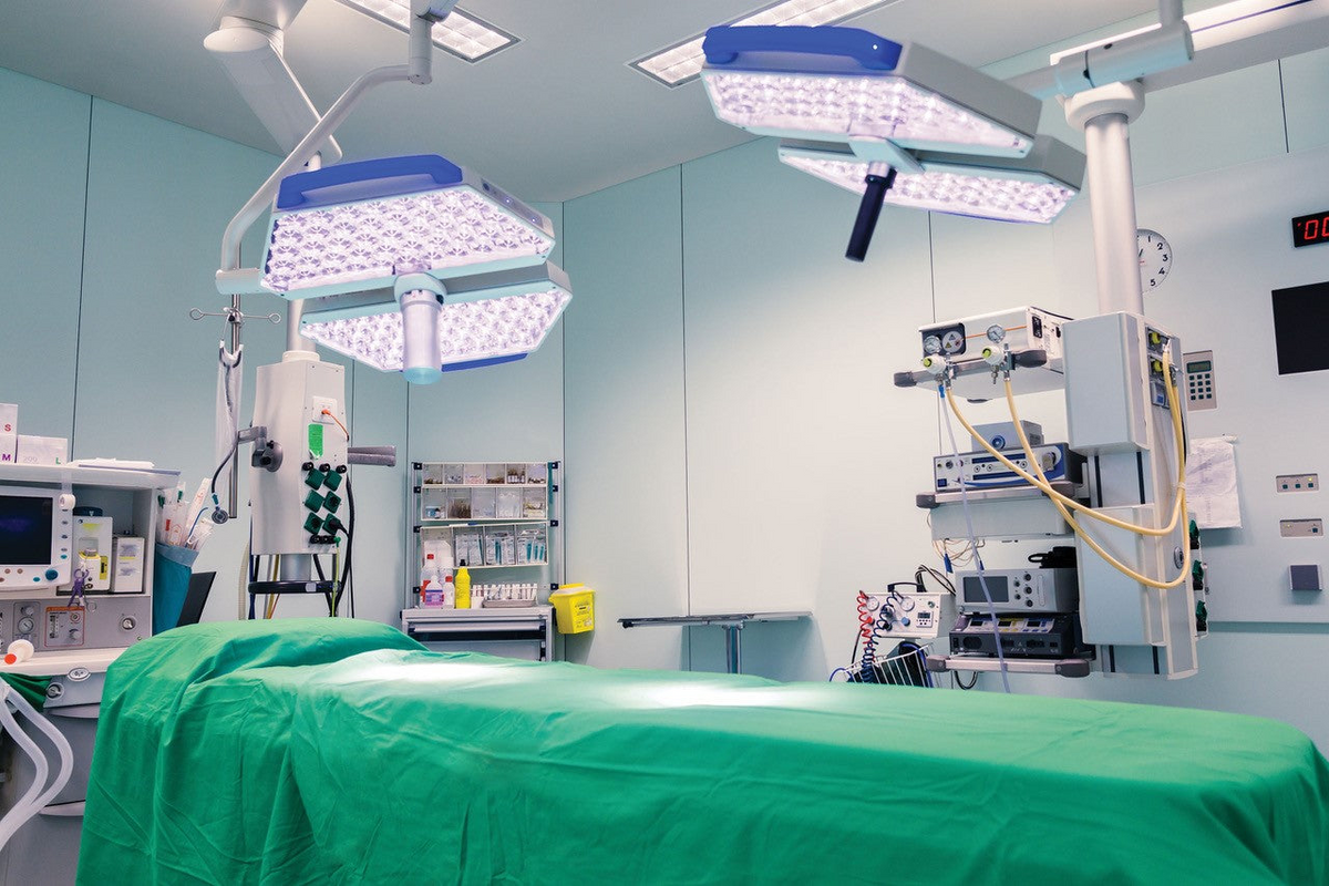 A sterile operating room features a surgical table draped in Thomaston Mills Healthcare Items - T-180 Jade Green, with bright lights, medical equipment, monitors, supply cabinets, and light blue walls.