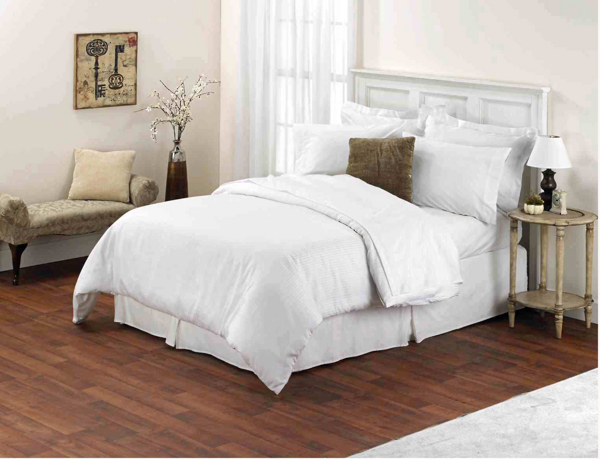 A neatly made bed features Thomaston Mills’ Pillowcases - Thomaston Brand Single Pick Percale, with a brown pillow in a bright room with wood floors, side table, lamp, bench with cushions and flowers, window, and framed artwork.
