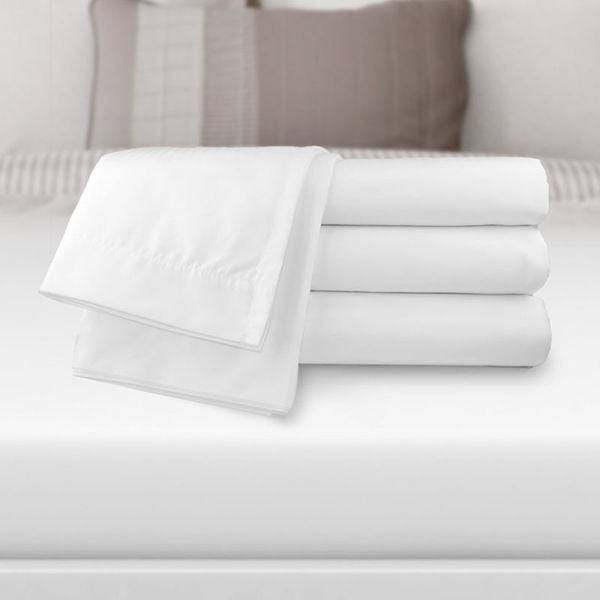 A set of Ganesh Mills Flat Sheets 2" Top Hem & 1" Bottom Hem - Oxford T200 Superblend Bed Linen is stacked on a bed with pillows and a headboard in the background, ideal for those seeking premium wholesale bedding.