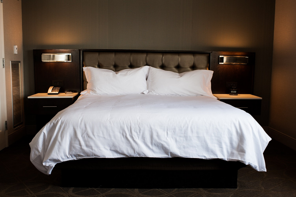 A neatly made bed with white linens and two pillows features Thomaston Mills Duvet Covers – 310 Thread Count Satin Stripe, centered between nightstands in a modern hotel room with a dark upholstered headboard.