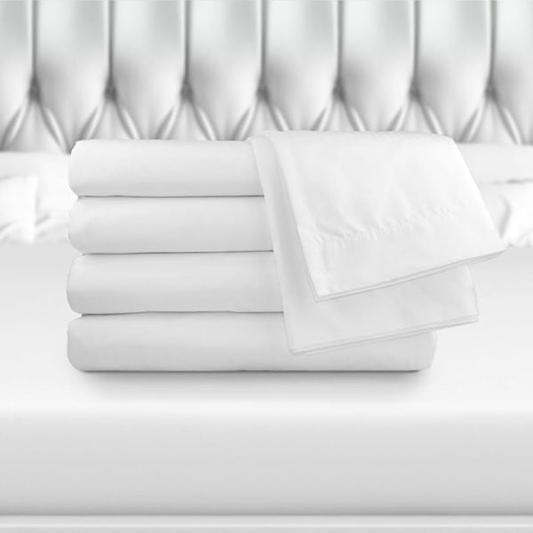 A stack of Ganesh Mills Oxford Super T300 Bed Linen fitted sheets is neatly folded on a white bed with a tufted, upholstered headboard in the background.