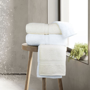 A stack of white and cream towels and the Ganesh Mills Bathmat - White/Ivory Oxford Vicenza sit on a wooden stool by a sunny window, with a small green plant on the sill.