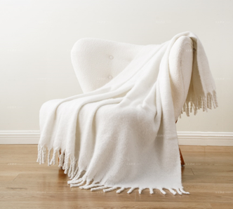The Pro Towels Vanilla Dream Throw, with chic fringed edges, is draped over a modern white upholstered chair against an off-white wall and light wood floor.
