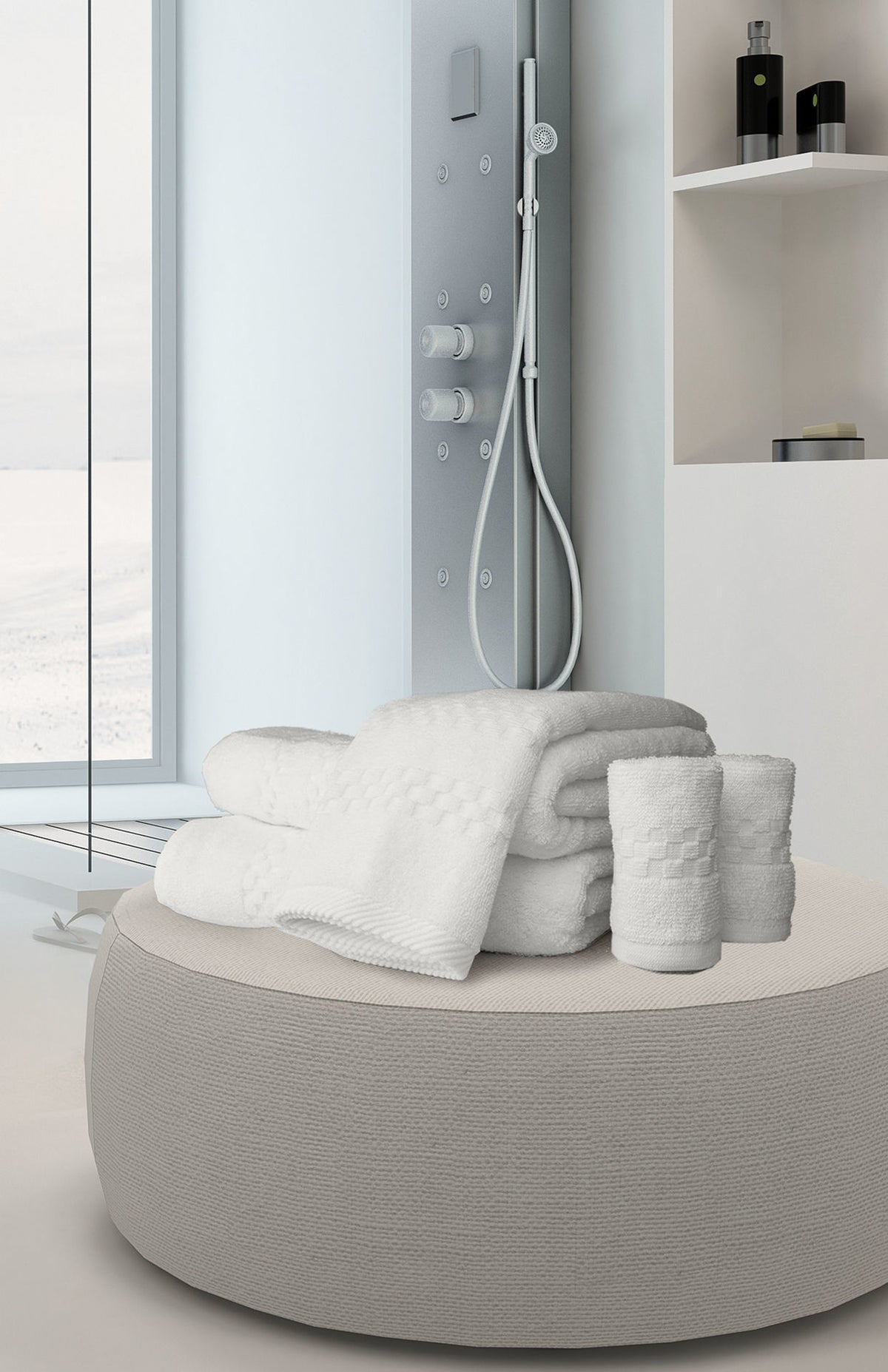 A stack of white Ganesh Mills Oxford Viceroy Bathmats and two rolled hand towels sit on a round ottoman in a modern bathroom with a glass shower and built-in shelves in the background.