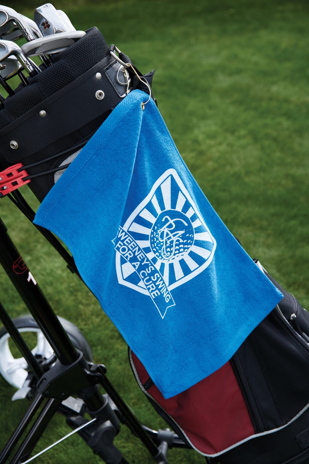 A blue Velour Fingertip Golf Towel by Pro Towels, featuring "WEEKEND SWING FOR A CURE," hangs from a golf bag on the course, standing out as a stylish promotional accessory against the green grass.
