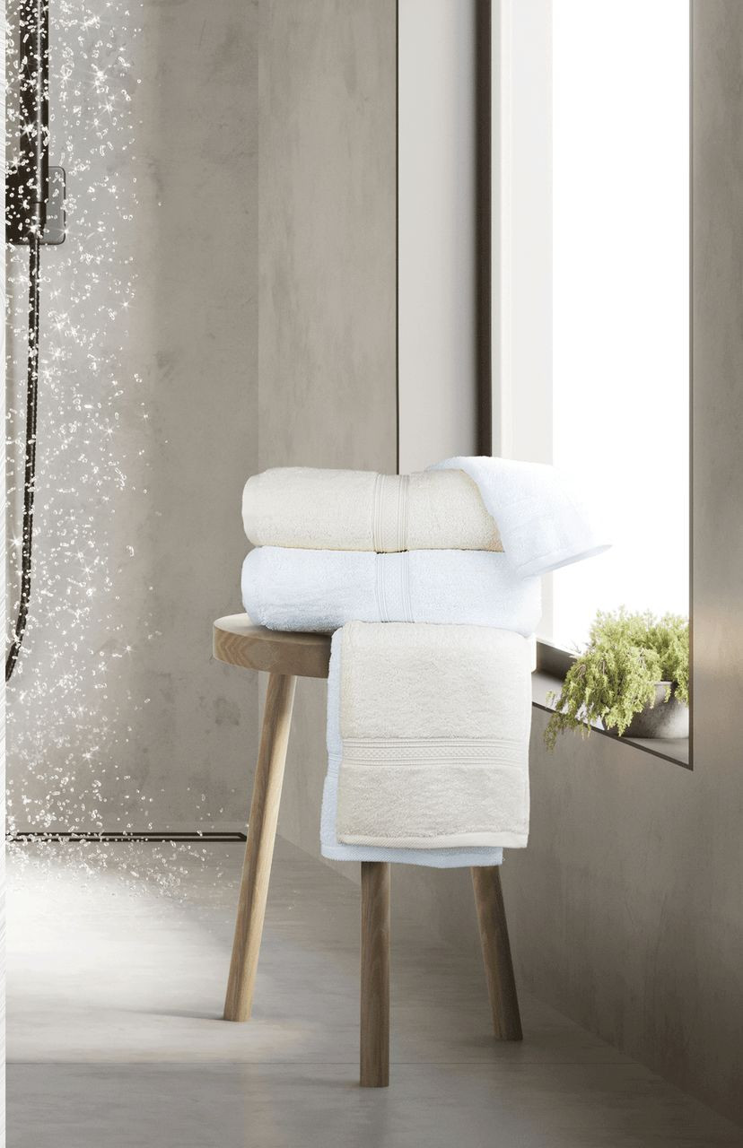 A stack of white towels and a Ganesh Mills Washcloth - White/Ivory Oxford Vicenza Towel rests on a wooden stool by a sunlit window, with greenery on the sill and water gently spraying in the shower background.