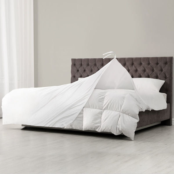 A neatly made bed with a tufted gray headboard and white bedding. The Ganesh Mills Heavy Duvet Insert - Baffle Box Design is easily lifted off the bed in one piece using a large hanger, highlighting its simple removal for cleaning.