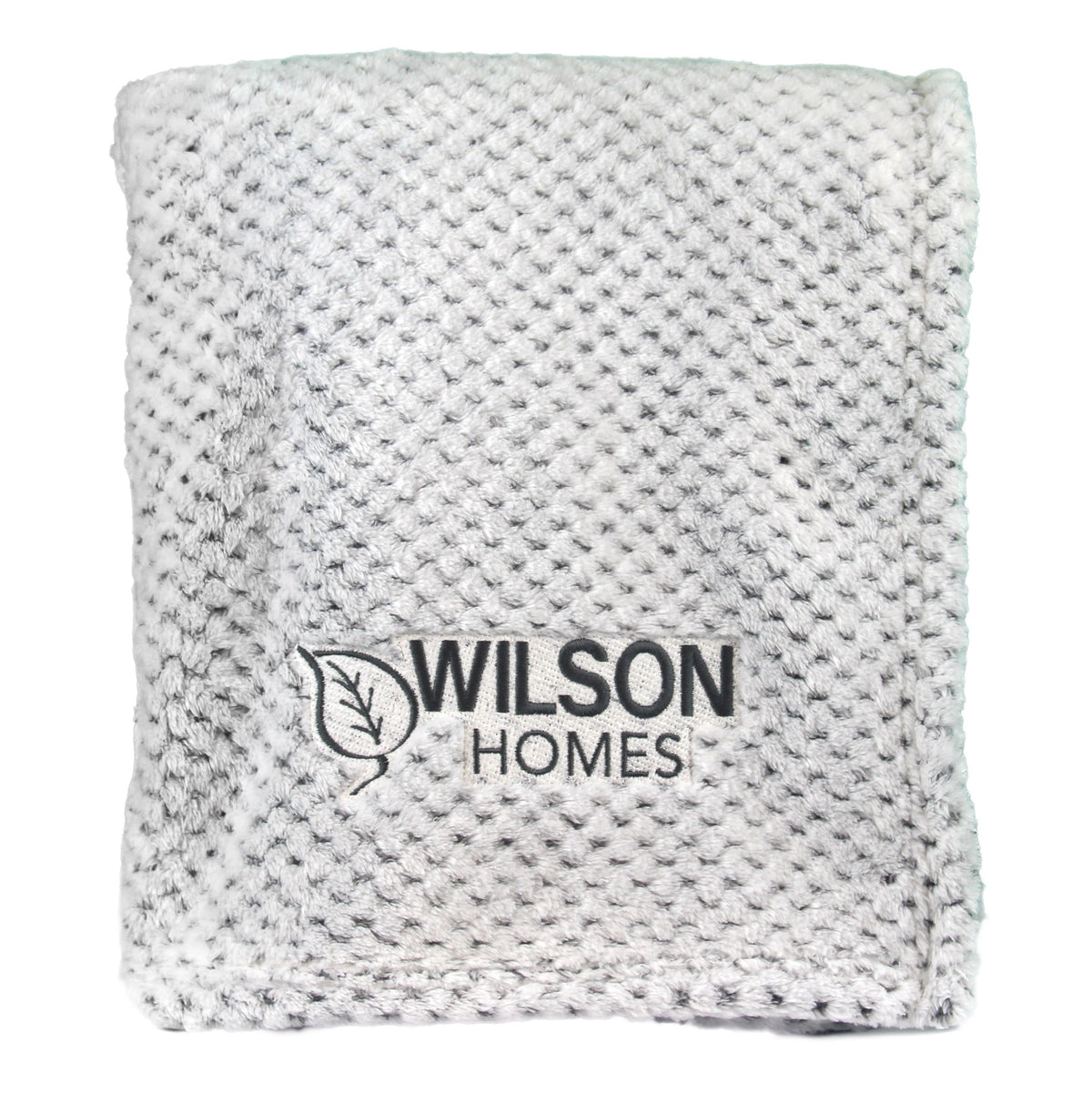 The Wanderlust Blanket by Pro Towels is a soft, white textured travel blanket, folded neatly with custom black Wilson Homes and leaf logo embroidery on the front.