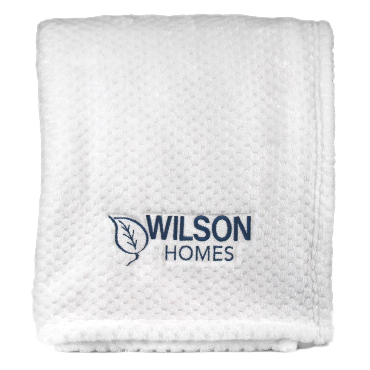 A folded white textured Wanderlust Blanket by Pro Towels, custom embroidered with the Wilson Homes name and leaf logo in blue thread on the front.