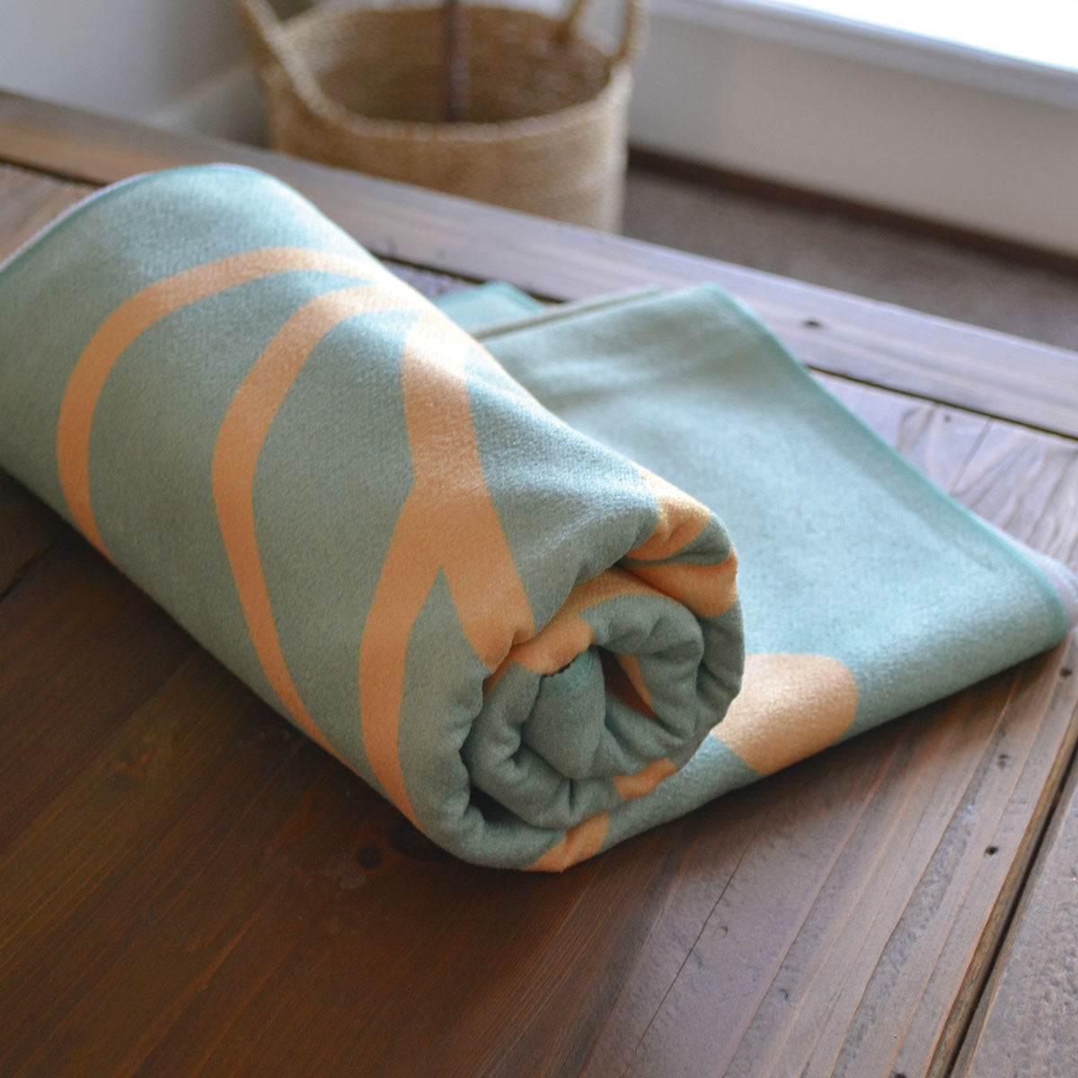 A rolled-up Pro Towels Yoga Mat Fitness Towel with a green and orange pattern sits on a wooden table, with a woven basket and window in the background.