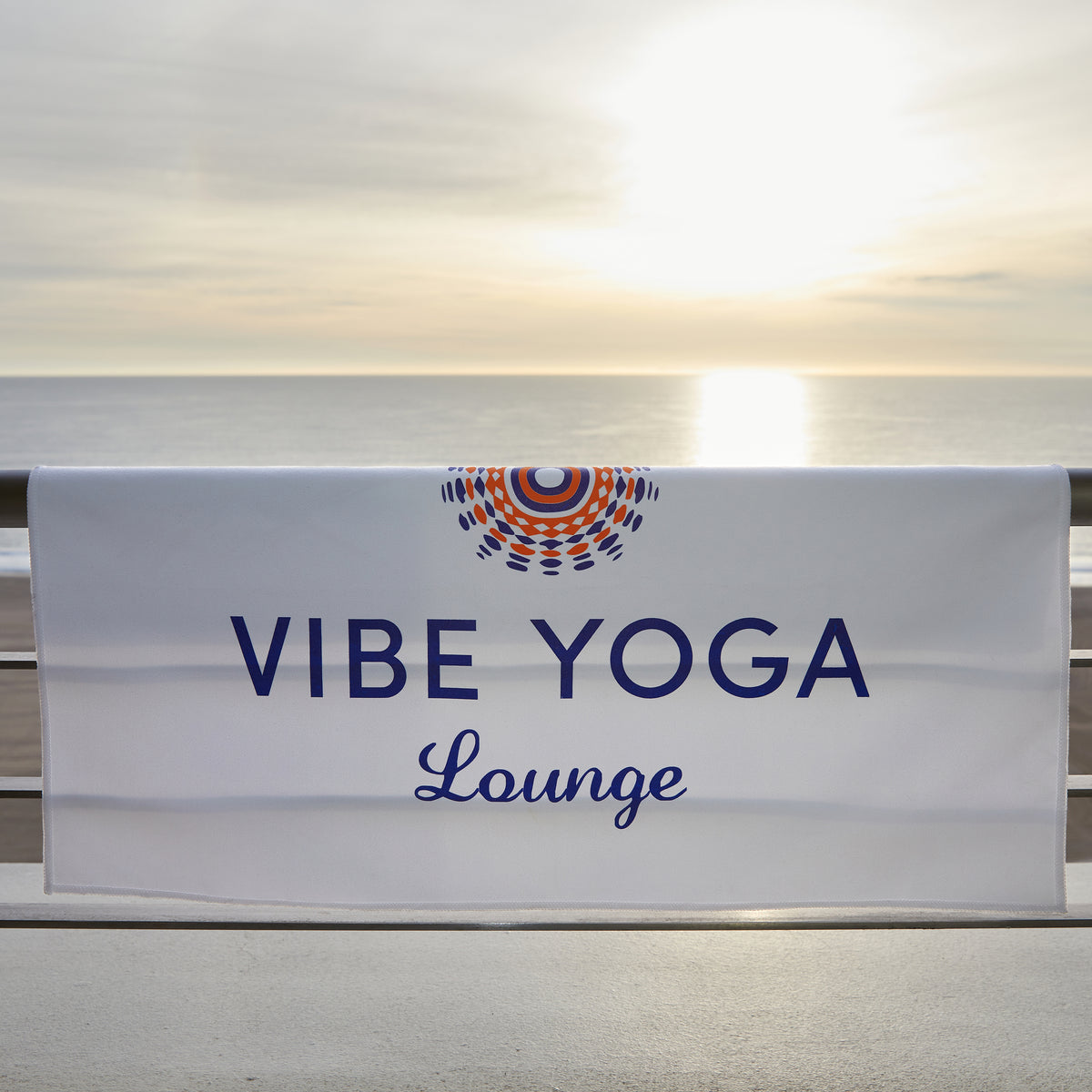 A white banner for Vibe Yoga Lounge hangs on a railing by the ocean at sunset, next to a Pro Towels Yoga Workout & Sports Towel—imprint-ready and ideal for your next beach yoga session.