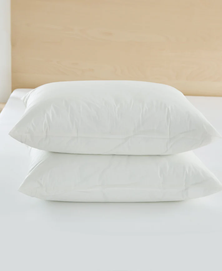 Two white pillows stacked on a white bed with a light wooden headboard, each encased in a Bargoose AllergyCare™ Cotton Allergen Barrier Pillow Protector for extra comfort and protection.