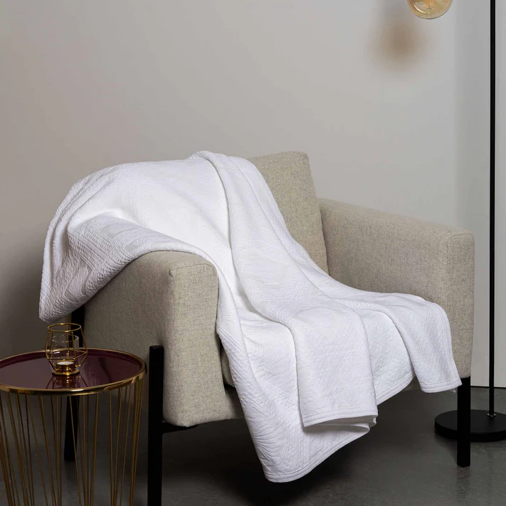 A light grey armchair is draped with an Avalon Jacquard Bedspread by Bargoose, beside a round side table holding a glass of water and a floor lamp with an exposed bulb, creating a modern, minimalistic look.