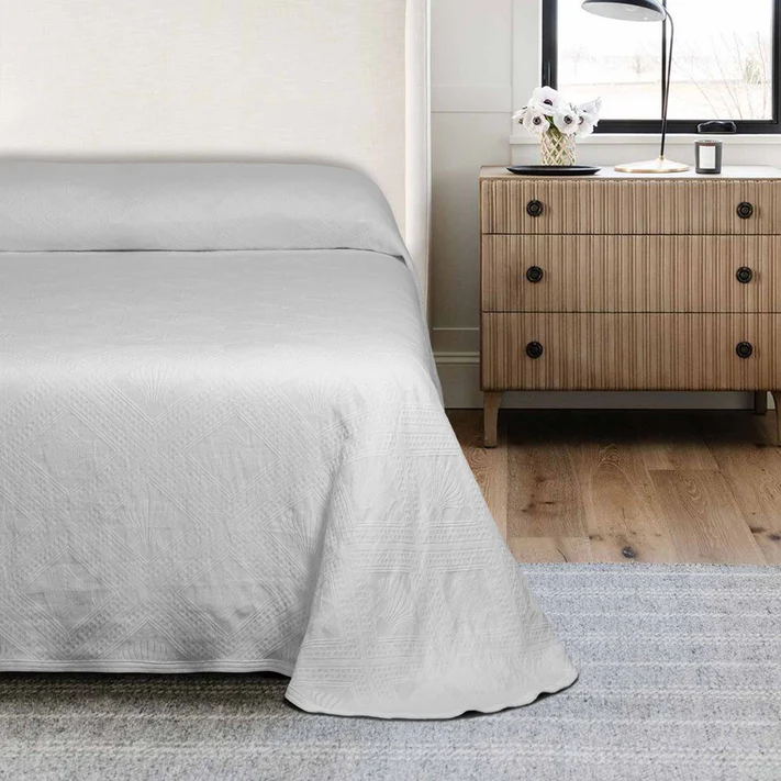 A bright, modern bedroom features a neatly made bed with the Avalon Jacquard Bedspread by Bargoose, next to a wooden nightstand with black knobs holding a vase of white flowers, atop a rug on wooden floors.