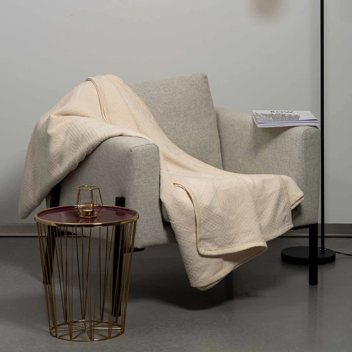 An Avalon Jacquard Bedspread by Bargoose is draped over a beige armchair beside a gold round side table with a glass. A magazine on the chair’s arm gives cozy, hotel-inspired vibes to this minimalist room with gray flooring.