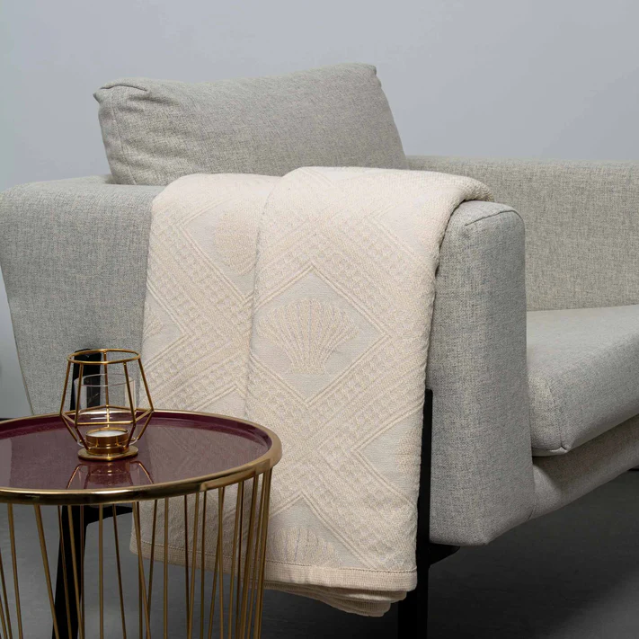 The Avalon Jacquard Bedspread by Bargoose is draped over a modern light gray armchair beside a round side table with a burgundy top and gold frame, which holds a geometric gold candle holder.