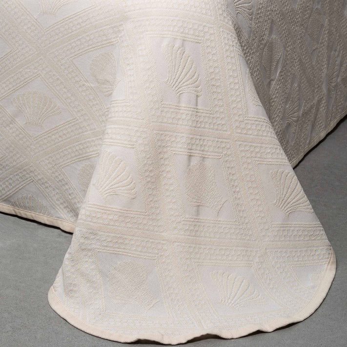 A close-up of the Avalon Jacquard Bedspread by Bargoose shows its cream color and detailed textured pattern with shells and geometric shapes, elegantly draped over a bed onto a gray floor.