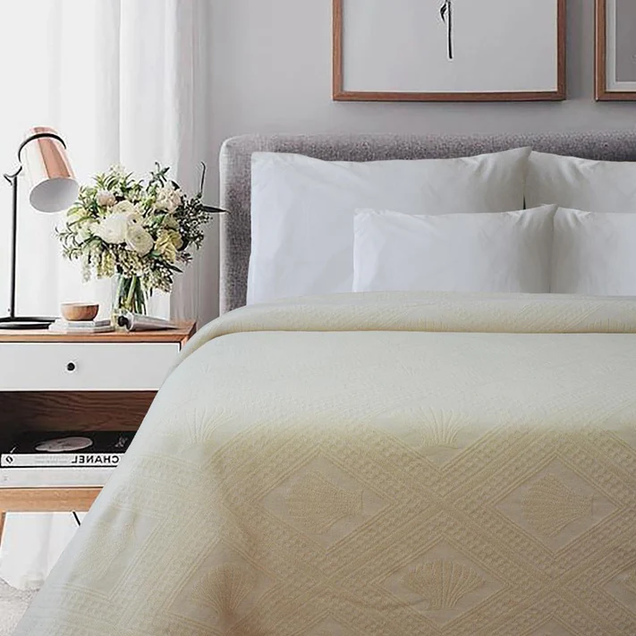 A neatly made bed features the Avalon Jacquard Bedspread by Bargoose, with white pillows. A bedside table holds a vase of flowers, lamp, and books beneath framed abstract art on a light wall, creating an inviting hotel bedding vibe.