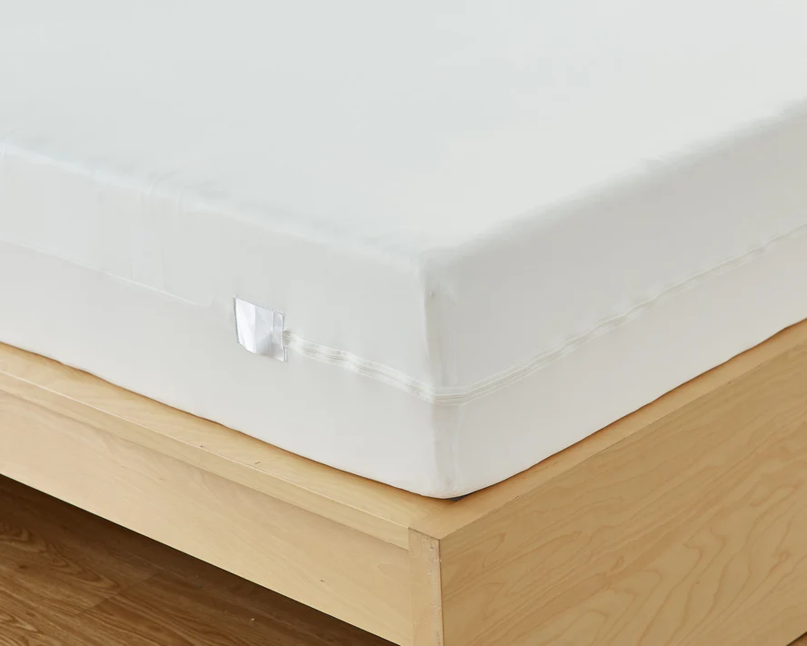 Close-up of a light wood bed frame with a white mattress, featuring the Bargoose 6 Gauge Vinyl Zippered Cover for mattresses up to 12 deep, and a small fabric tag on the side.